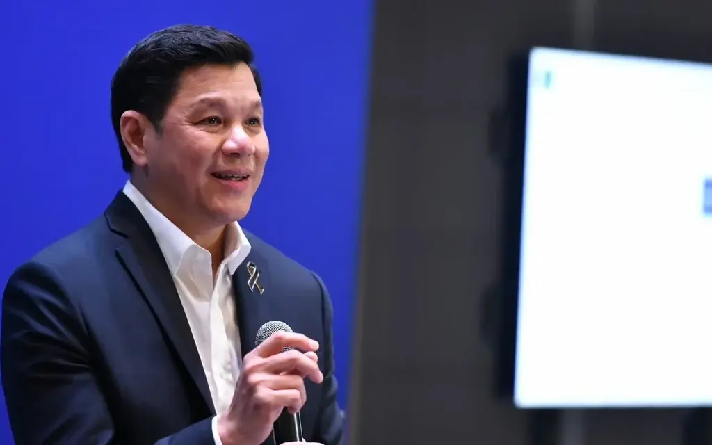 A suited man speaks into a microphone beside a presentation screen.